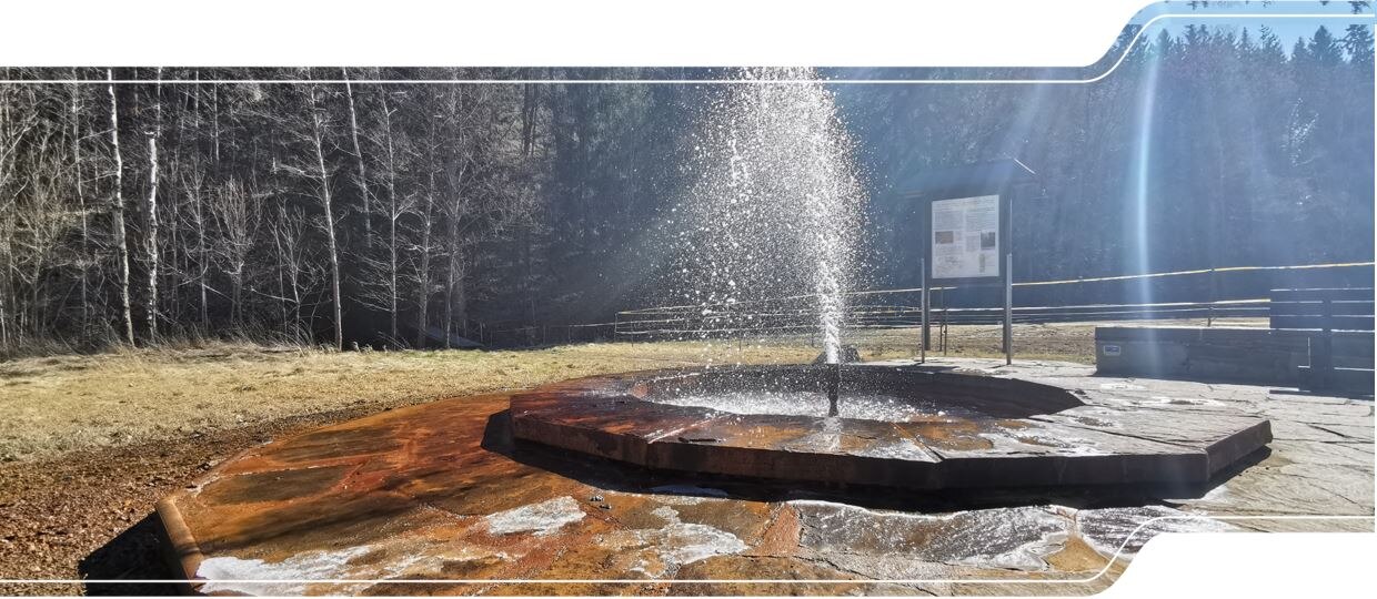 Mit Naturstein eingefasster Springbrunnen mit Erklärungstafel daneben.