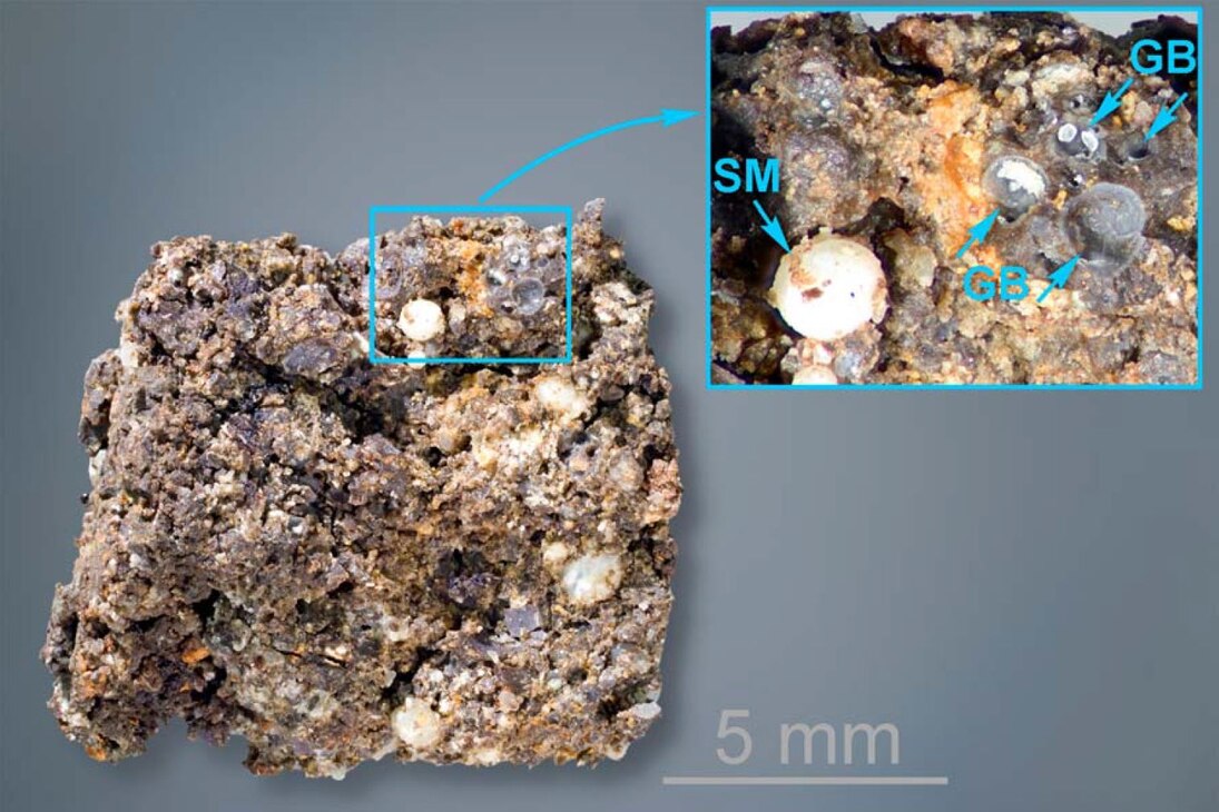 Basaltische Schlacke mit Gasblasen (GB) sowie kugeligen Ausfüllungen der Gasblasen durch Sekundärminerale (SM). 