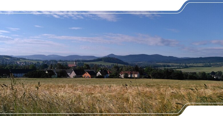 Feld mit Dorf im Flachland, dahinter waldiges Bergland